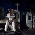 Merida Cementerio General Mexico Dia de los muertos white customed man with cowboy hat with skull make-up sitting on gravetomb surrounded by tombs at at the Festival Des Las Animas