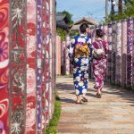 Japan Kyoto two women dressed in kimonos walk through poles with traditional colourful kimono fabrics at Arashiyama station
